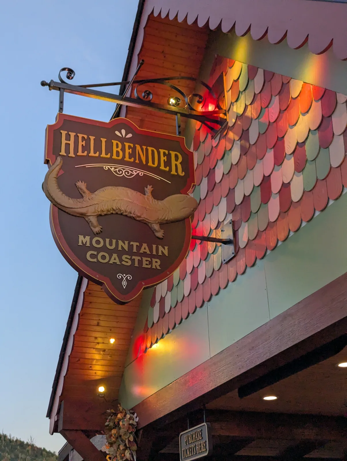 A sign reading Hellbender with Mountain Coaster underneath, lit at dusk, and empty track curving away through the trees