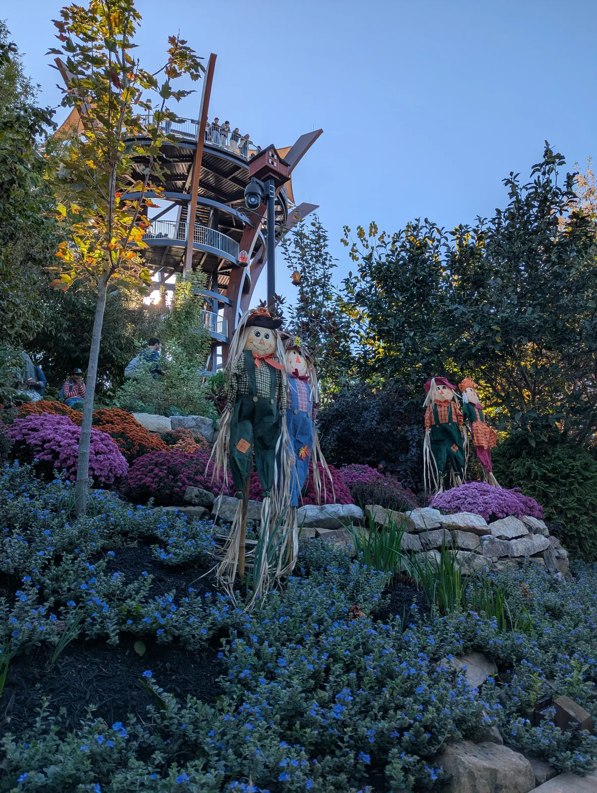 A scarecrow in a straw hat standing among red, yellow, and orange flowers, with a valley behind