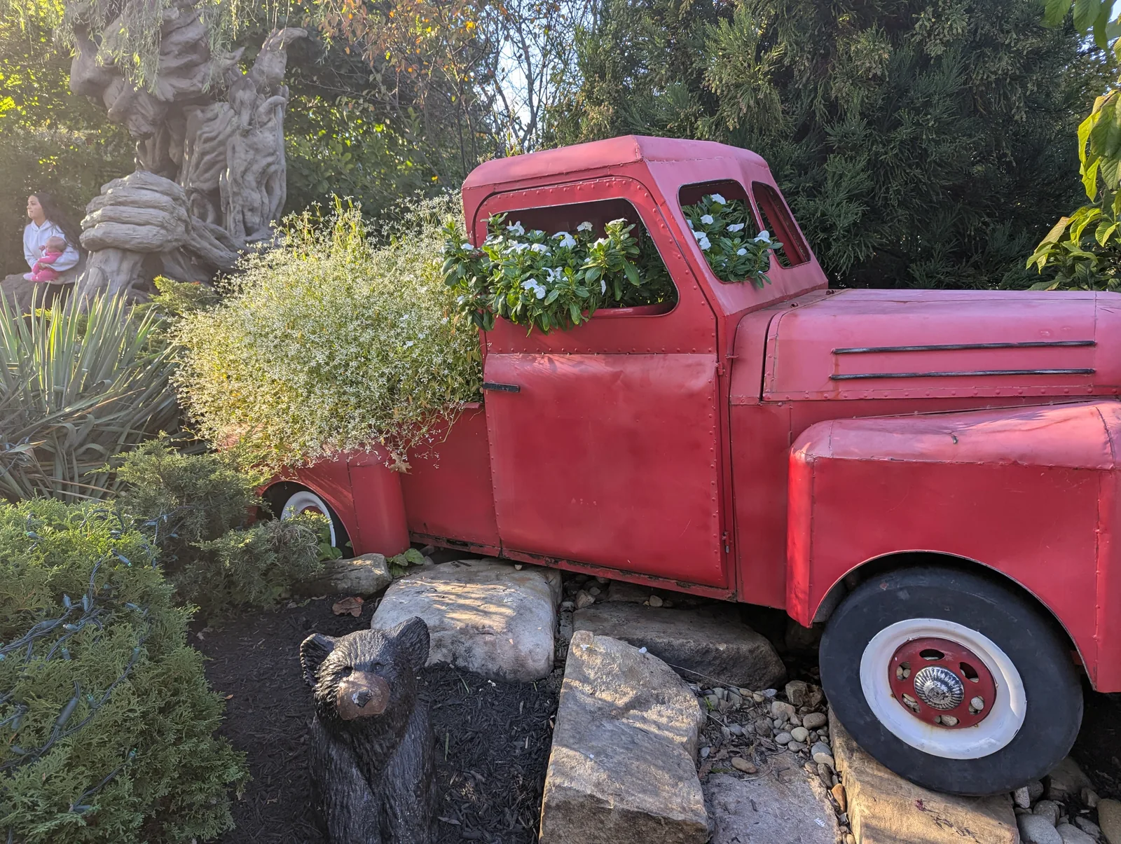 A vintage red truck with whitewall tires parked by a low stone wall, fall trees and a ridge behind it