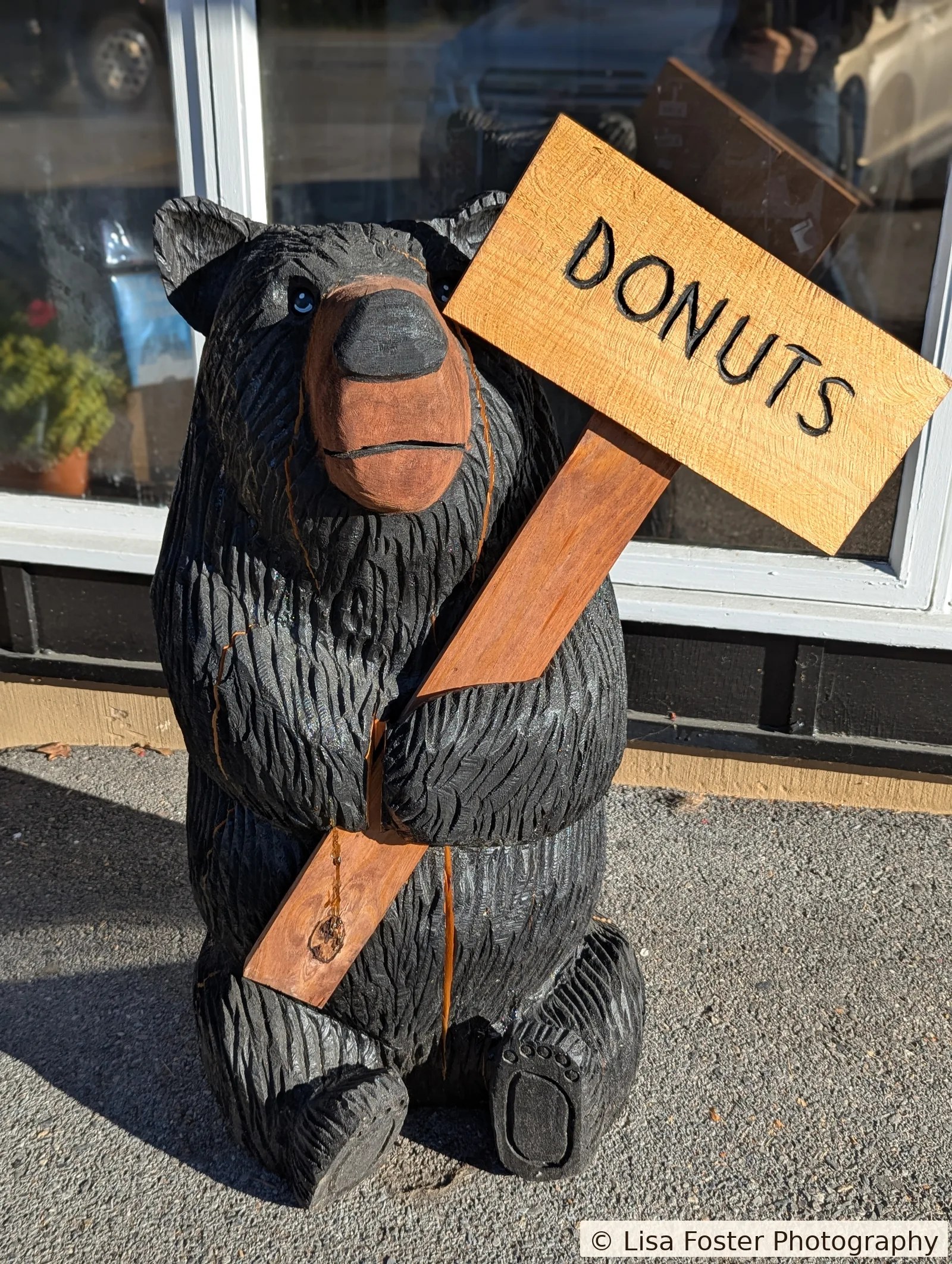 A carved wooden bear outside The Apple House holds a wooden sign that reads DONUTS, with fall trees behind it.