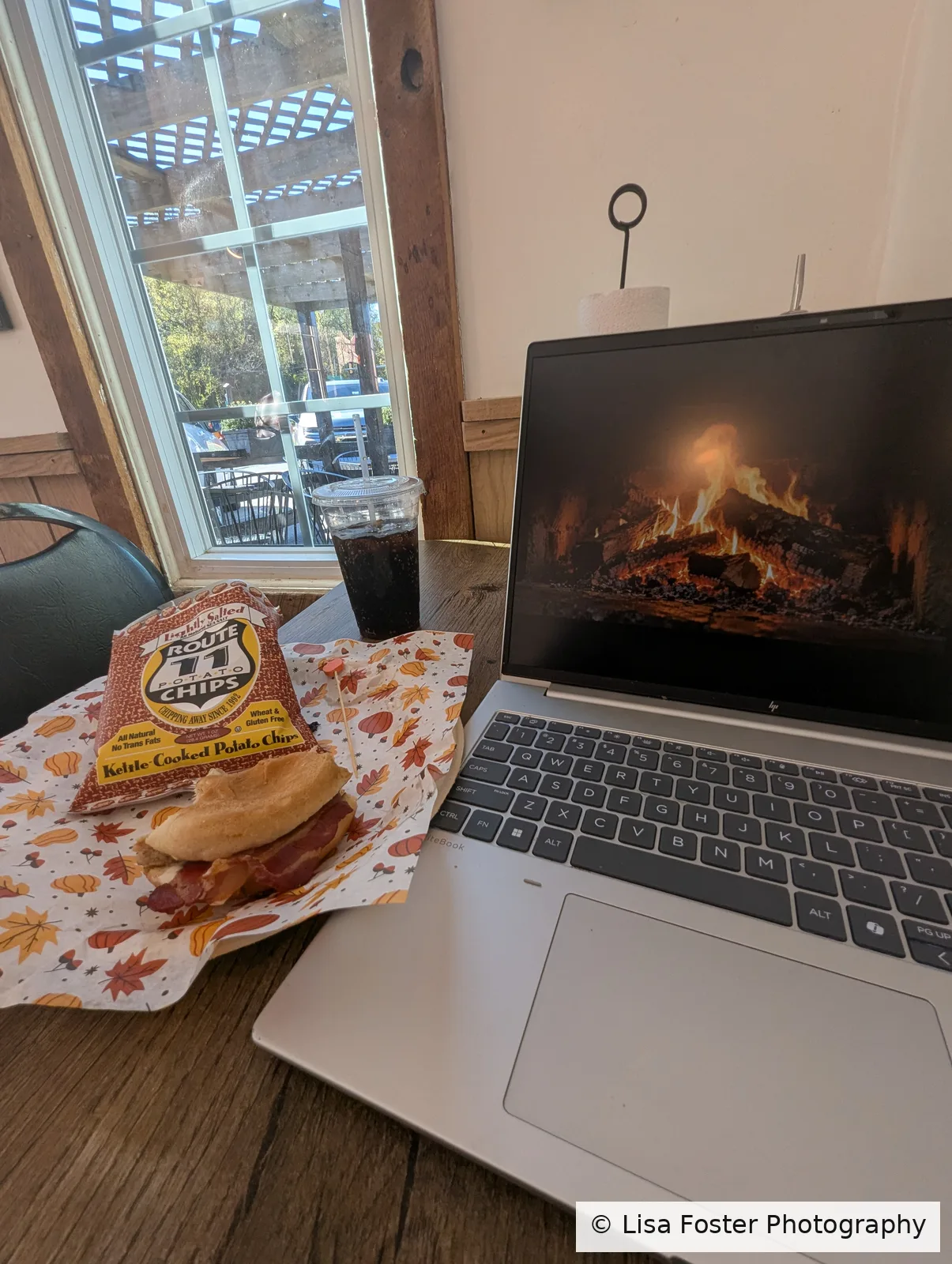 An HP laptop open on a table beside a window, with the Rooster (a grilled chicken sandwich), Route 11 chips, and a soda.