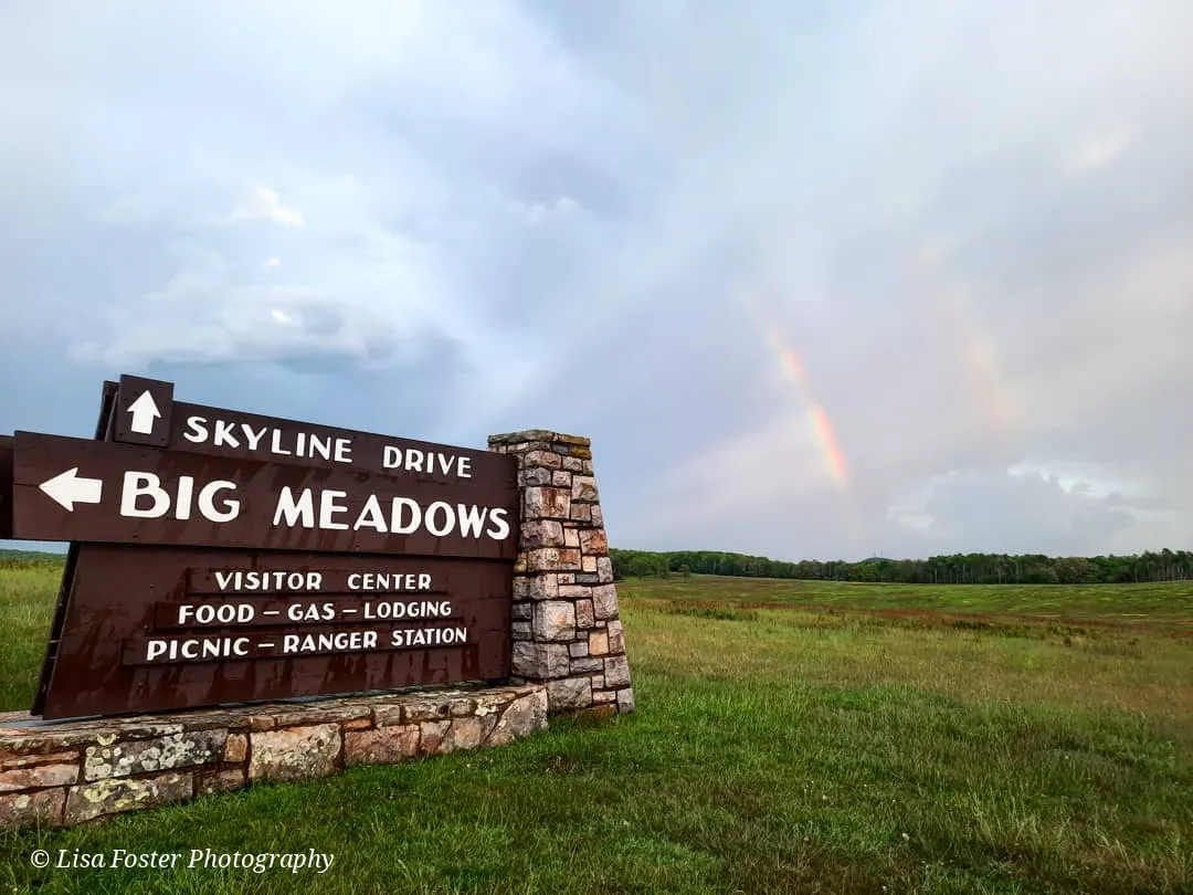 The stone and wood Big Meadows sign on Skyline Drive with a faint double rainbow over the green meadow behind it