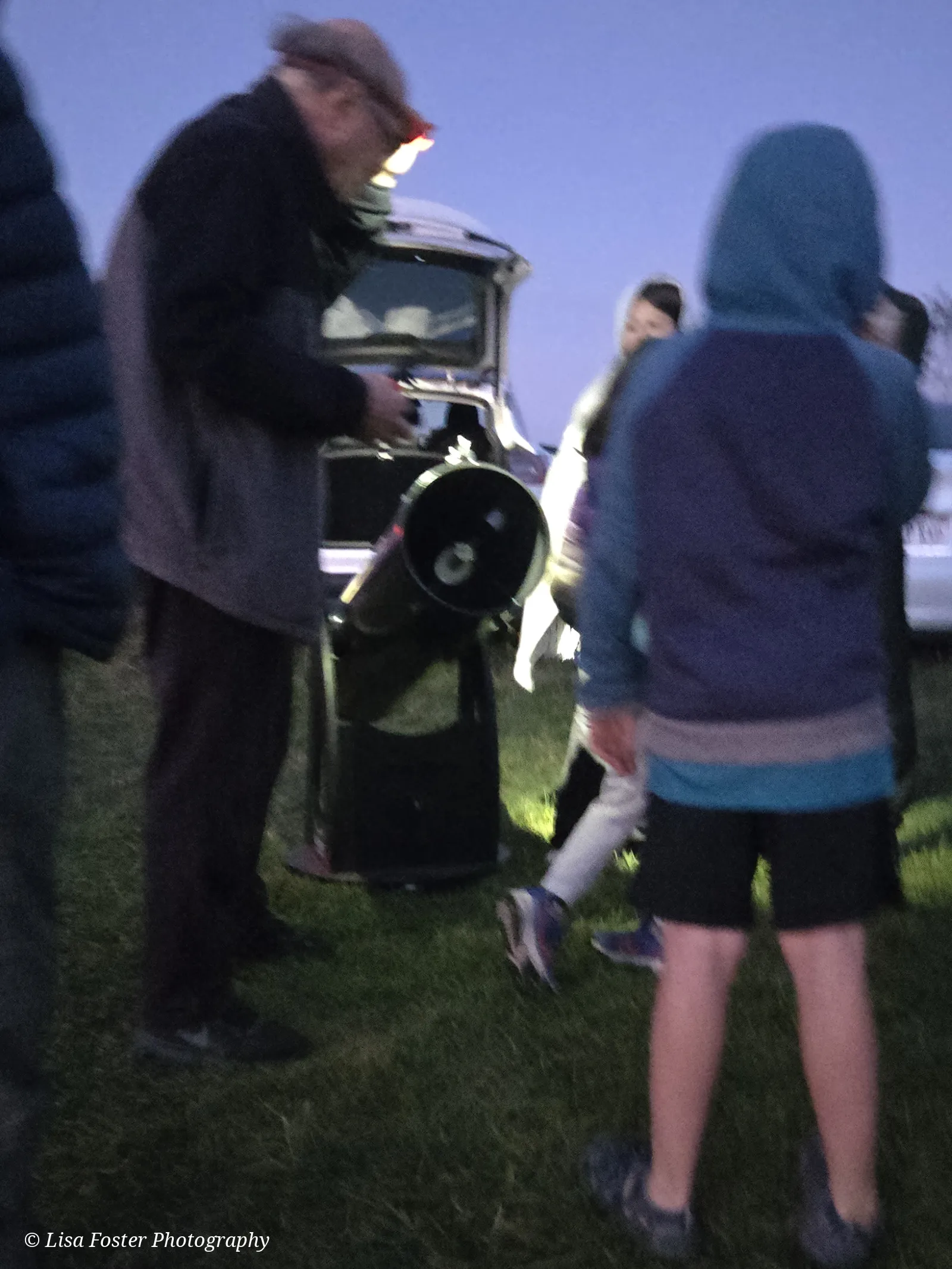 People in jackets standing on the grass around a large telescope at dusk
