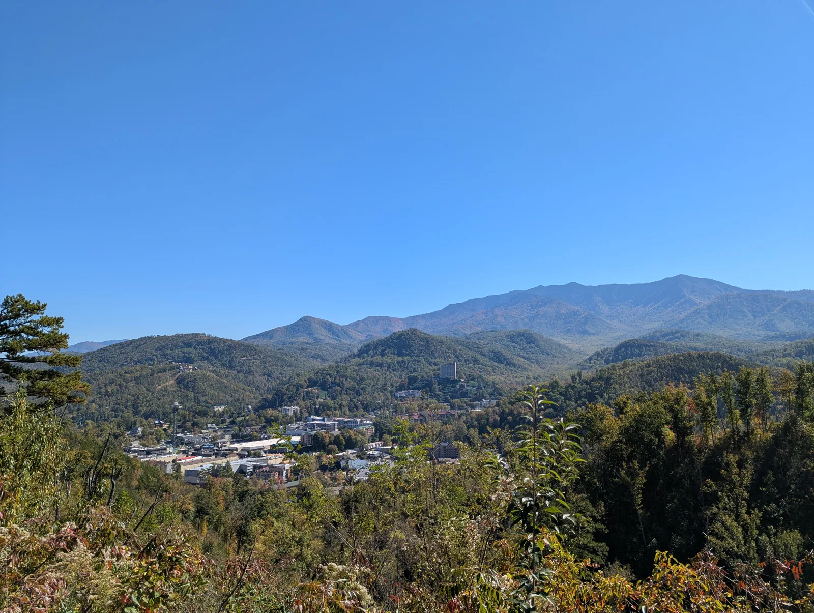 View from the Gatlinburg Bypass overlook: downtown Gatlinburg in a wooded valley, a tall high-rise among the buildings, the main Smoky Mountains ridge behind under a clear blue sky, and early fall color in the foreground trees