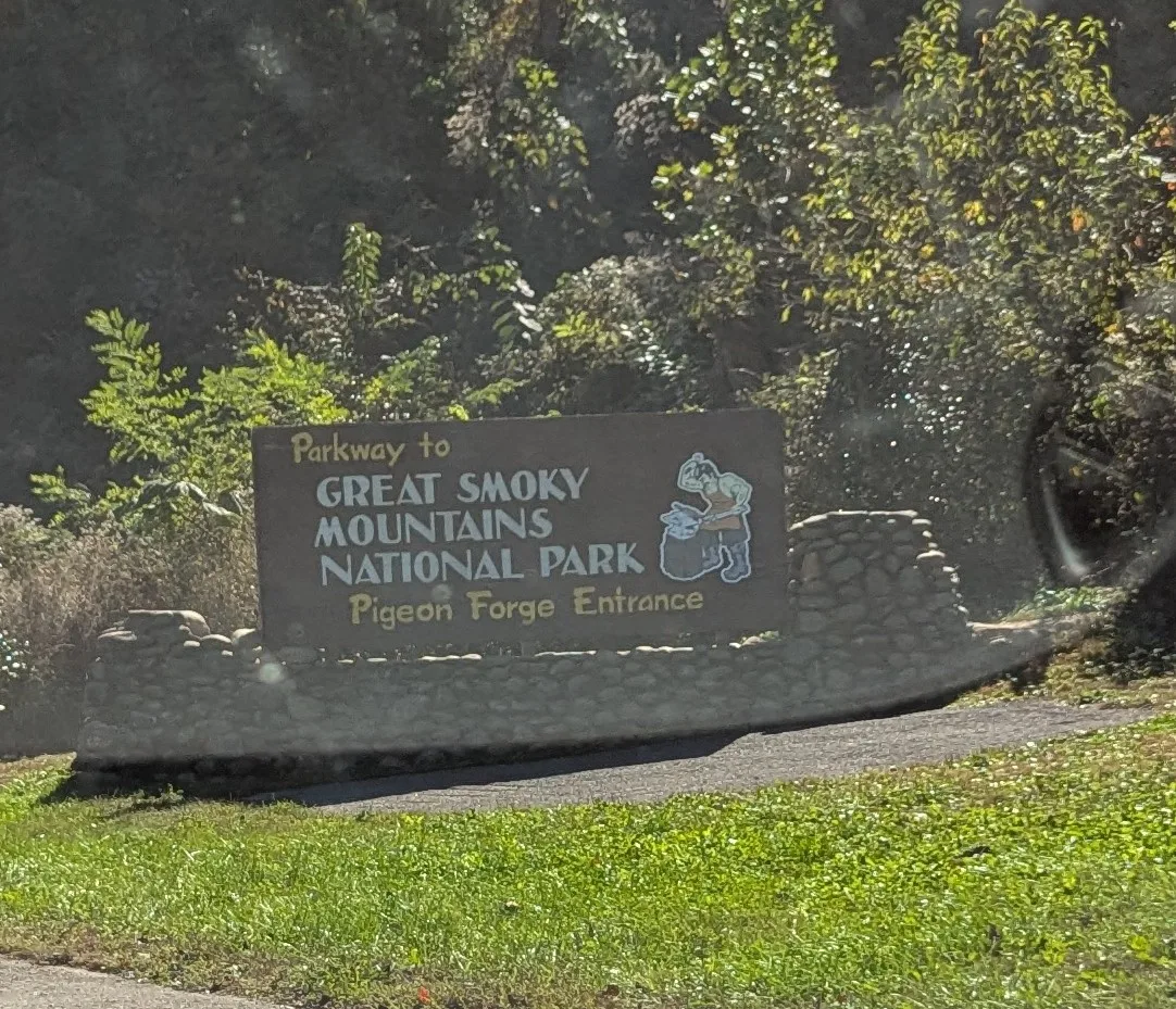 A stone sign reading "Parkway to Great Smoky Mountains National Park, Pigeon Forge Entrance" beside green shrubs