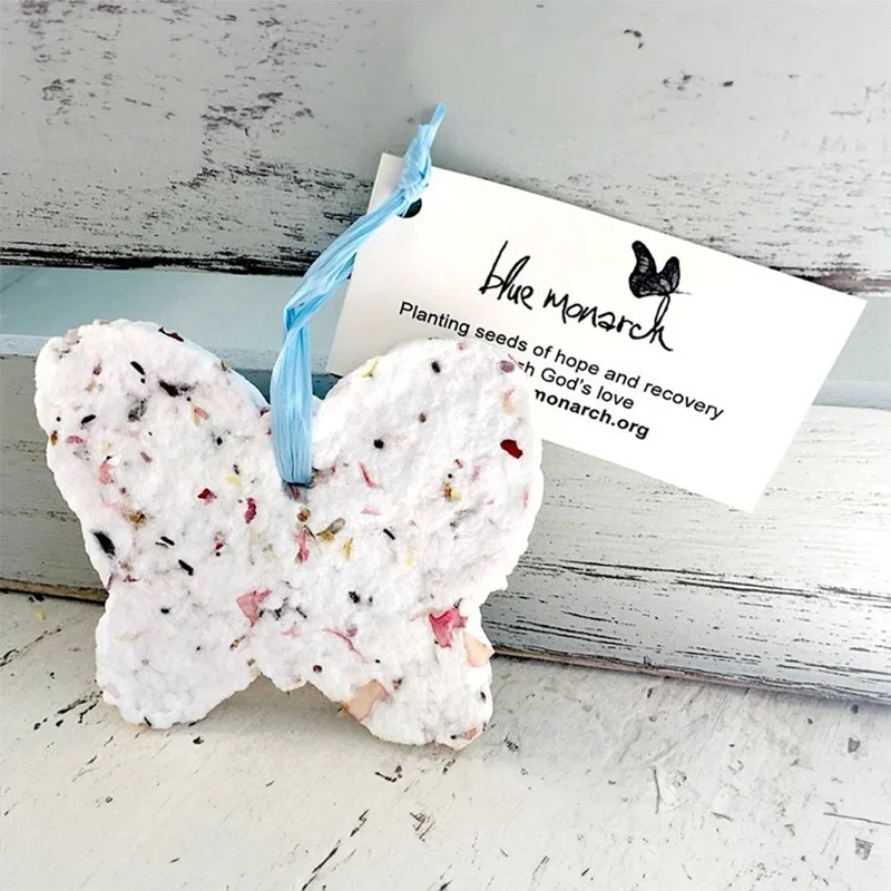 A butterfly-shaped seed ornament of recycled cotton fiber with pressed flowers, hanging from a blue ribbon.