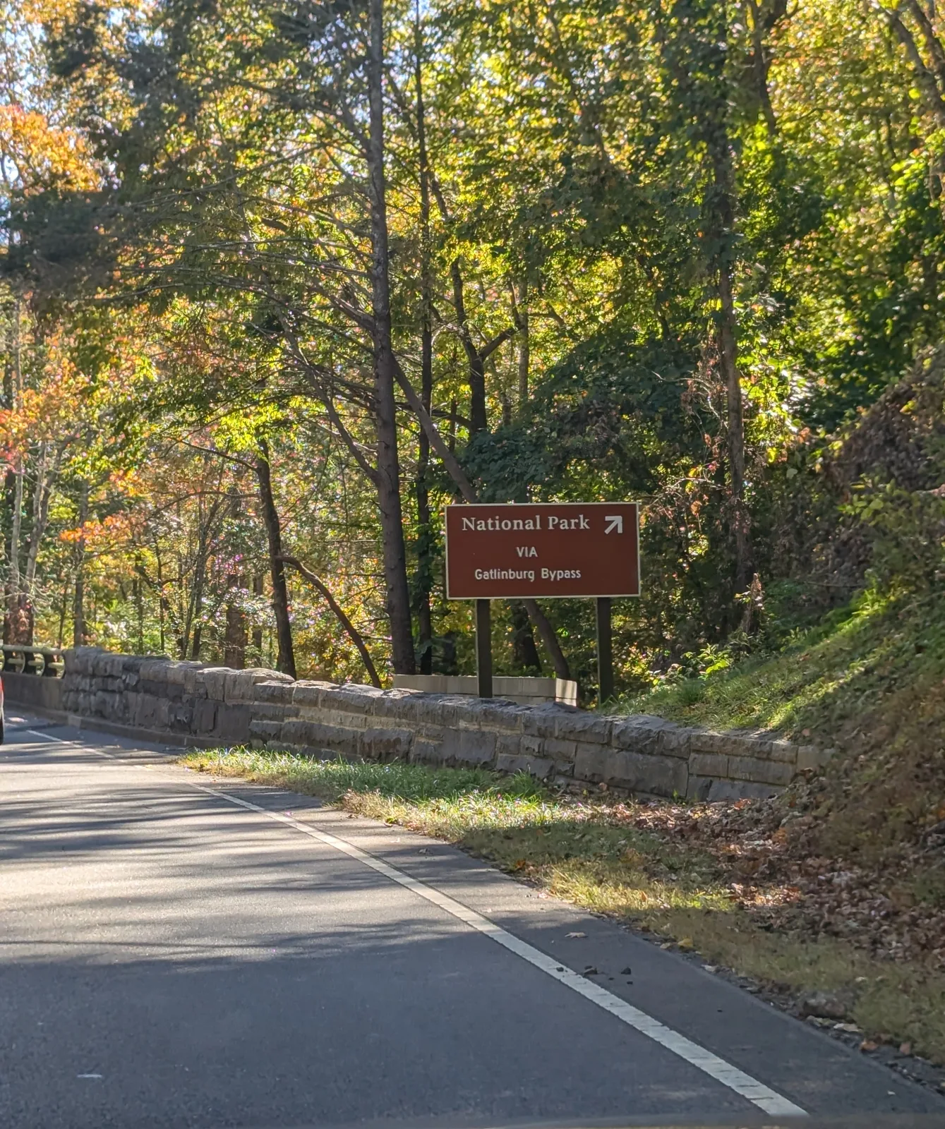 A brown "National Park via Gatlinburg Bypass" sign beside a stone wall, with trees in early fall color along the road