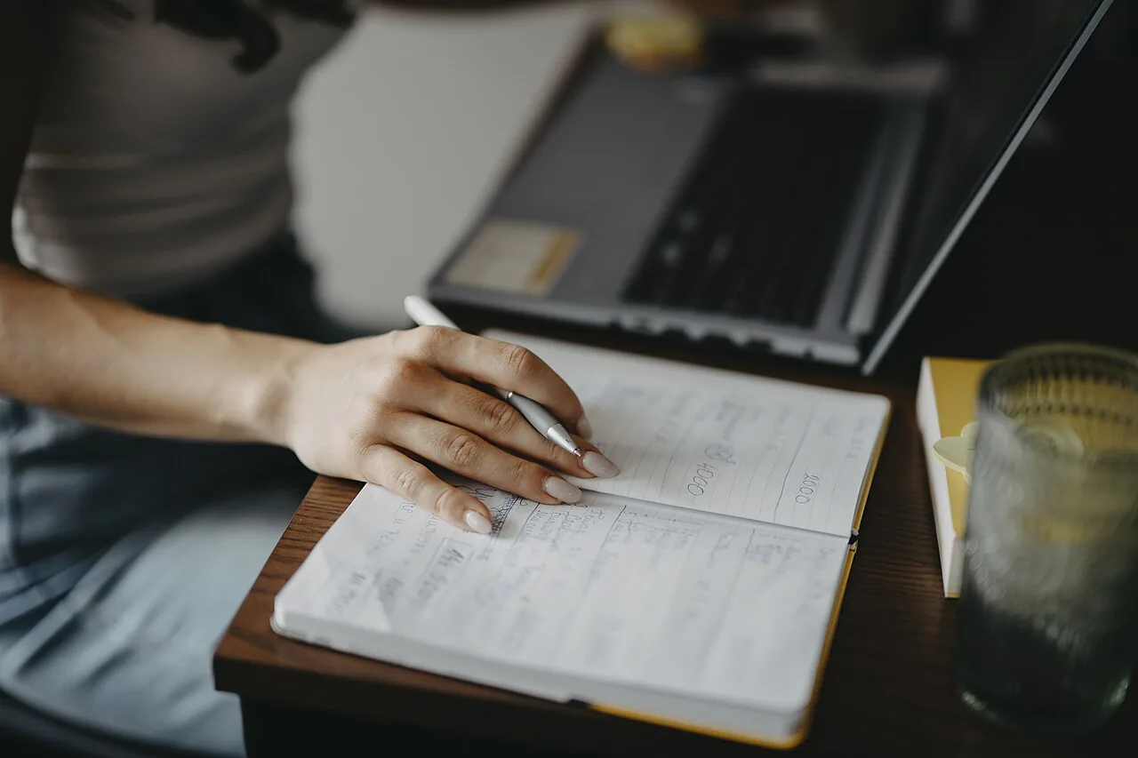 A person writing in a notebook next to an open laptop