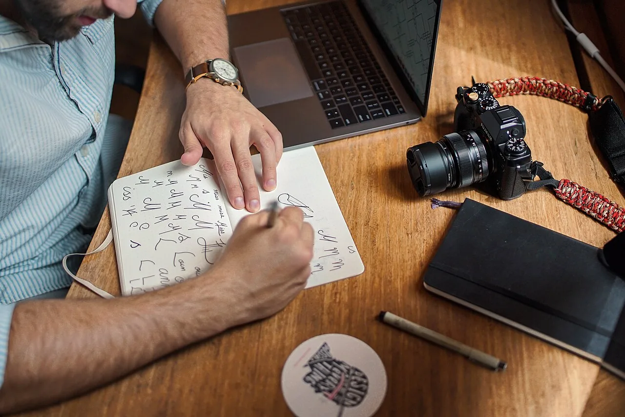 A person writing notes in a small notebook beside a laptop and a camera on a wooden desk