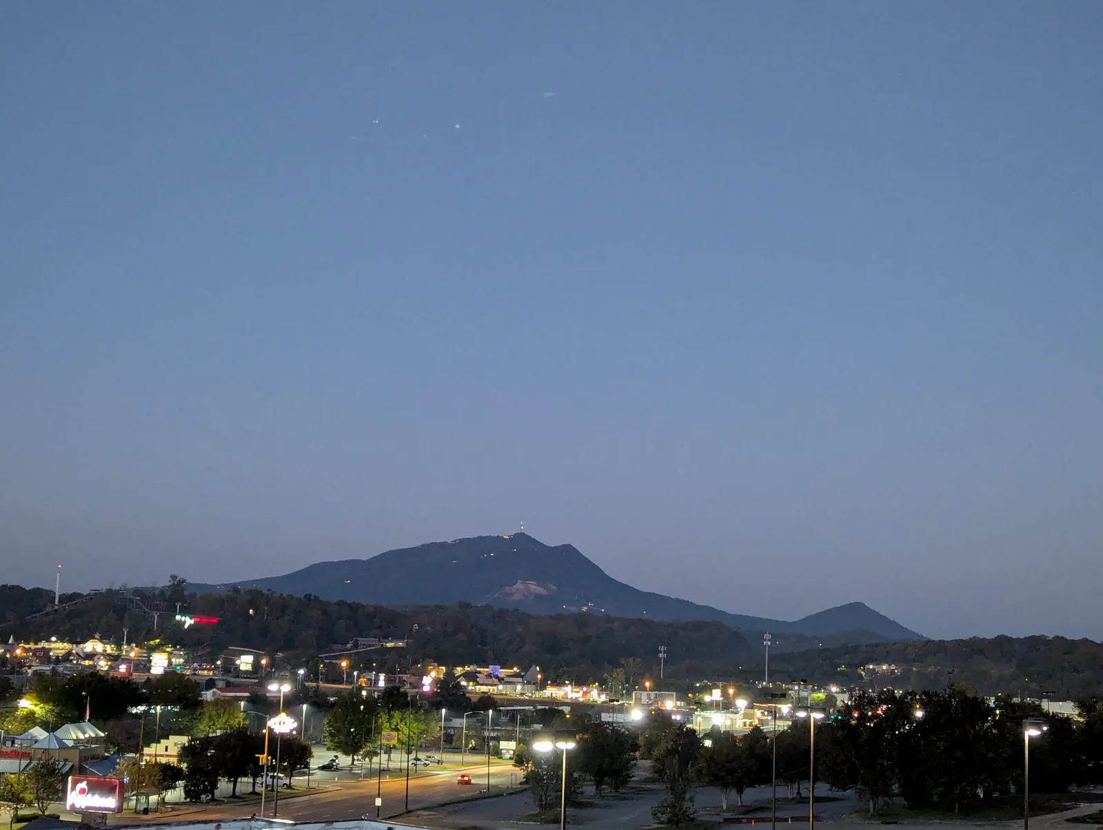 The sun in the upper left of the frame over a blue ridge, with rooftops and trees in the foreground under a pale sky