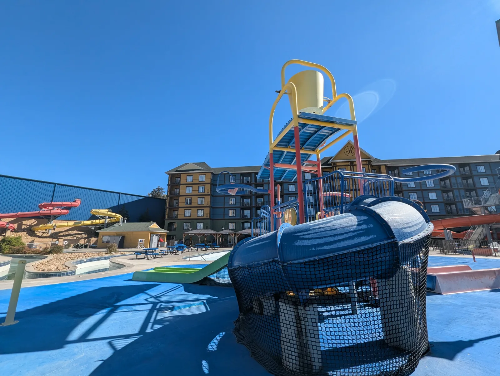An outdoor kids' splash structure with a yellow tipping bucket and blue slides, with resort buildings and red and yellow water slides behind it