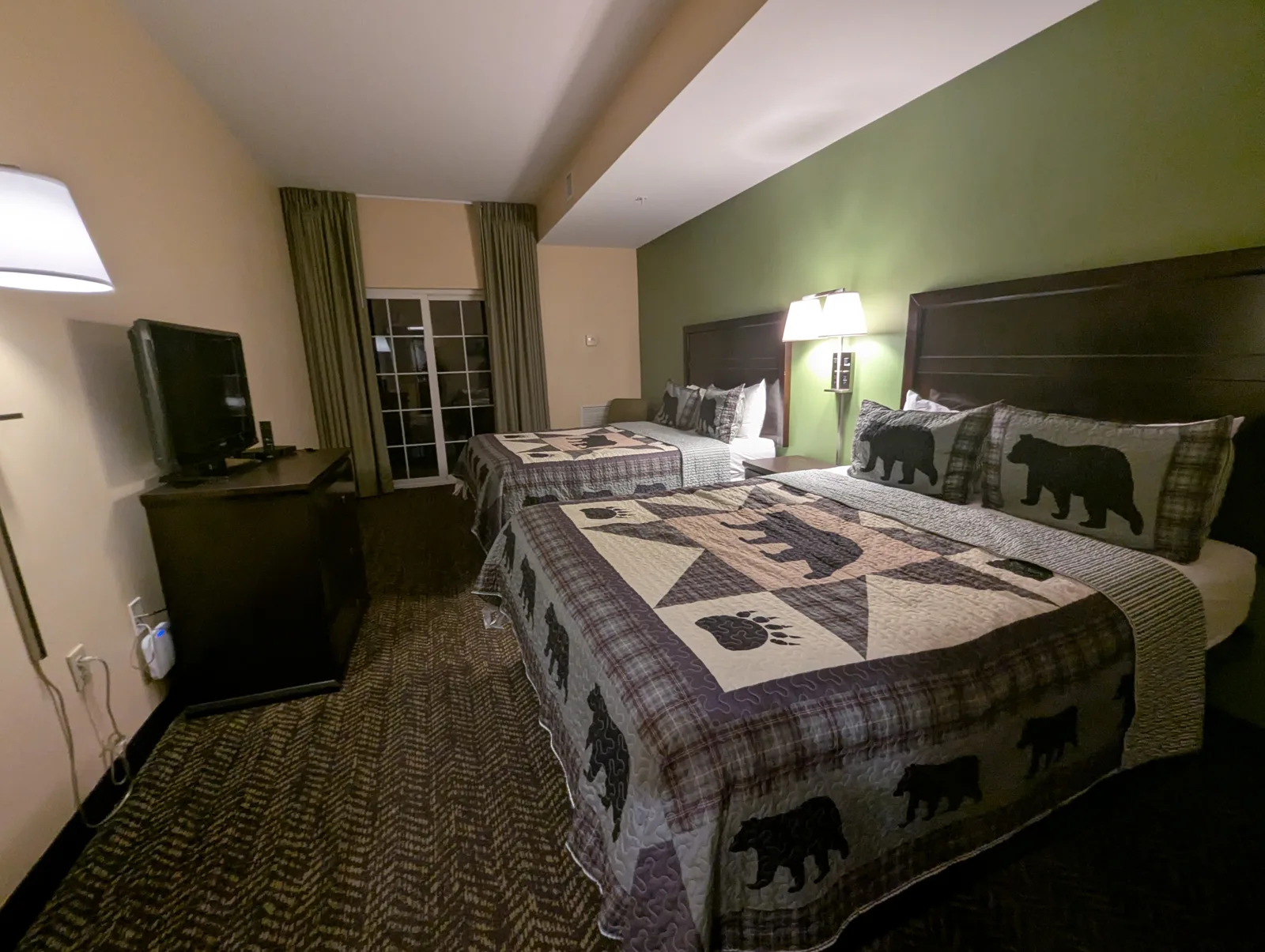 Two queen beds in the room with plaid quilts and pillow shams printed with black bears, a TV on a dresser, and a glass balcony door with long curtains
