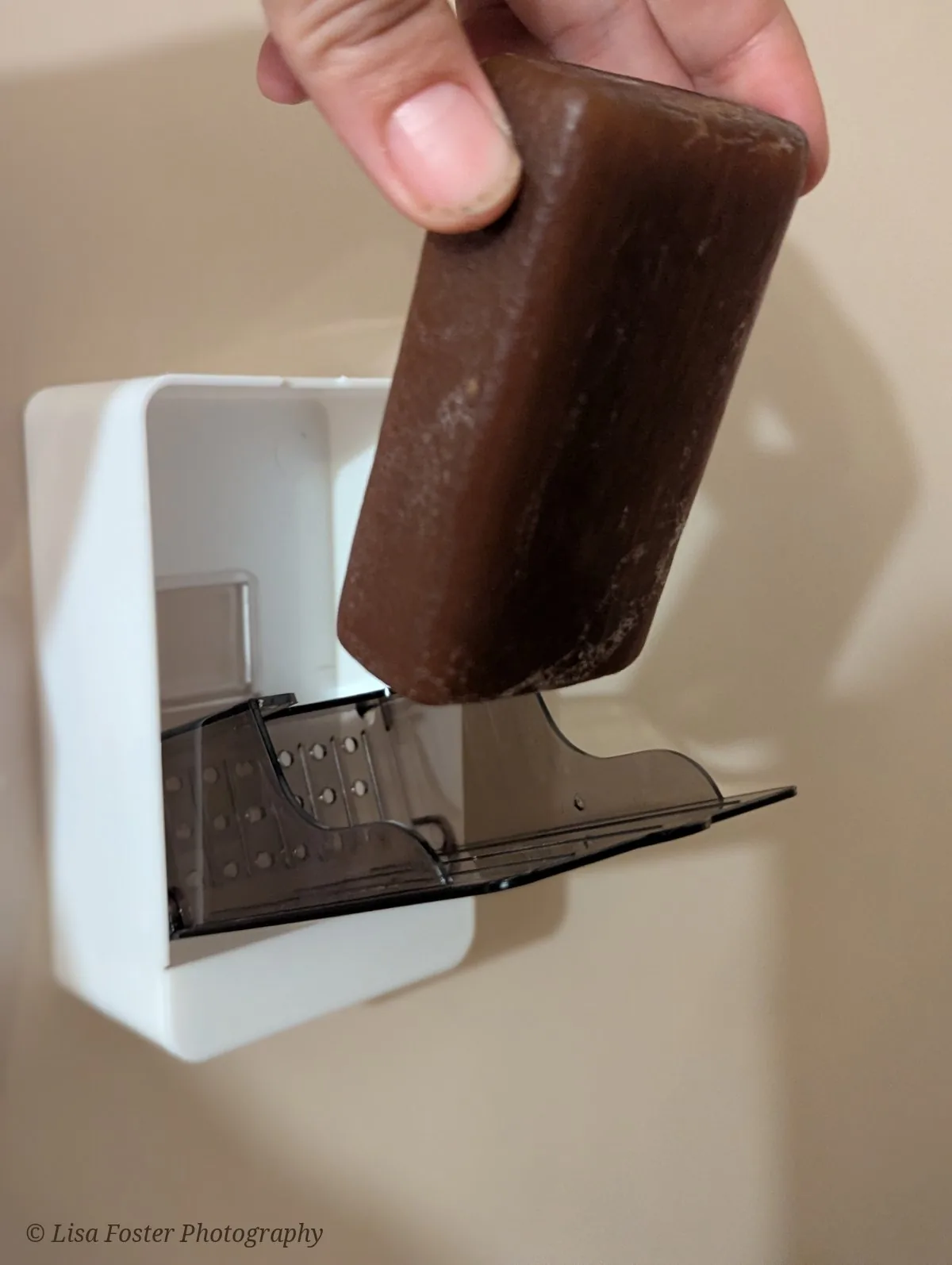 A hand lifts a dense, dark brown rectangular bar of birch tar soap out of a white wall-mounted soap holder