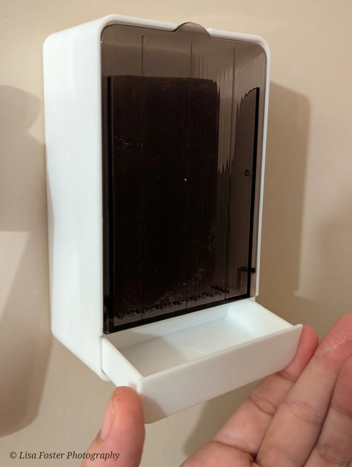 A hand pulls out the small white drip drawer at the bottom of the wall-mounted soap box