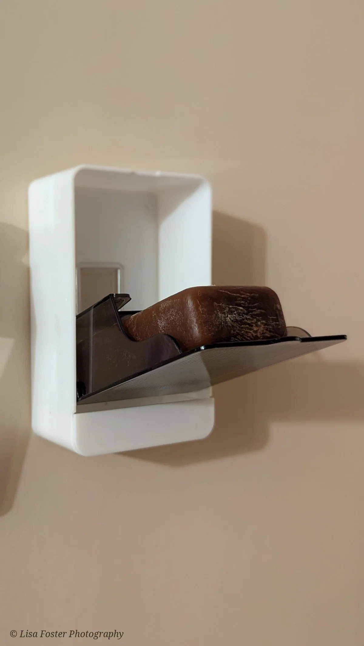 A dark brown bar of birch tar soap resting on the smoky flip-down tray of a white wall-mounted soap box